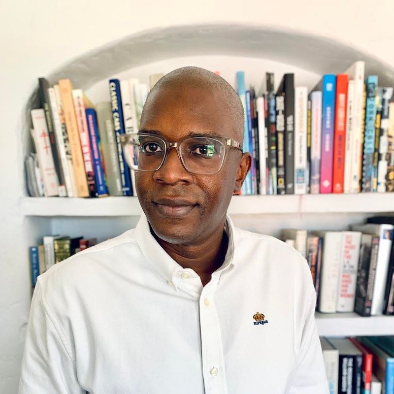 Patrick Kigongo (he/him) looking confidently at the camera with a bookcase behind him